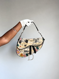 A Dior Saddle bag with a vintage Parisian illustration print, held by a hand wearing a white glove against a plain light gray background.