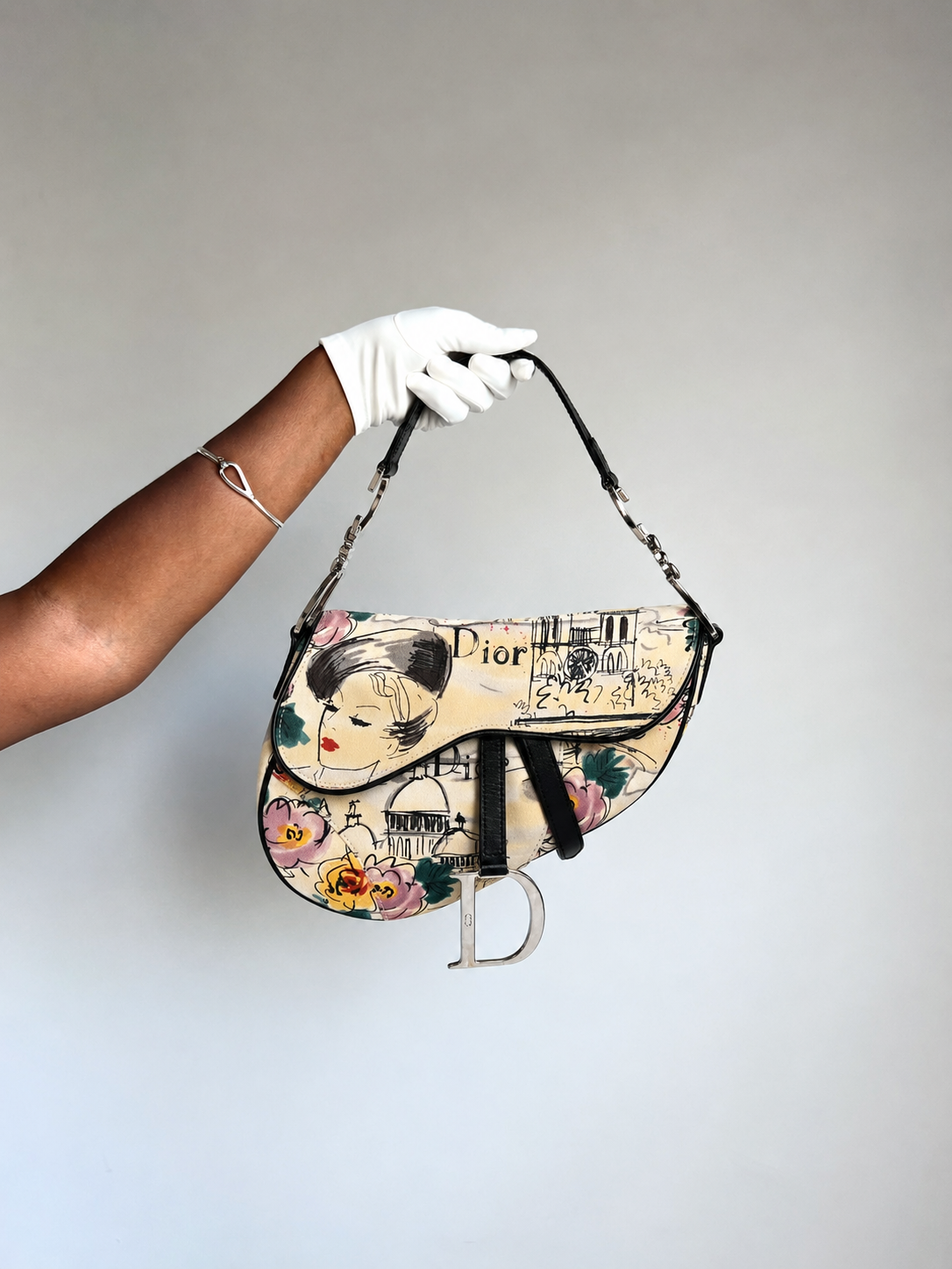 A Dior Saddle bag with a vintage Parisian illustration print, held by a hand wearing a white glove against a plain light gray background.