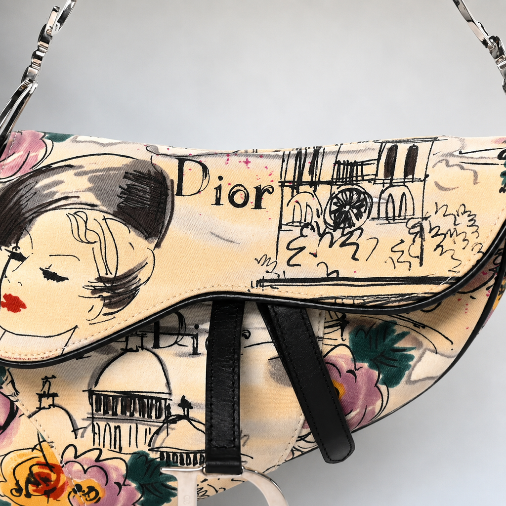 A Dior saddle shoulder bag featuring a cream-colored base printed with black sketch-style illustrations of a woman's face, the word 'Dior', Parisian landmarks, and pink, green, and yellow floral motifs. It has black leather strap accents and a silver-tone chain strap.