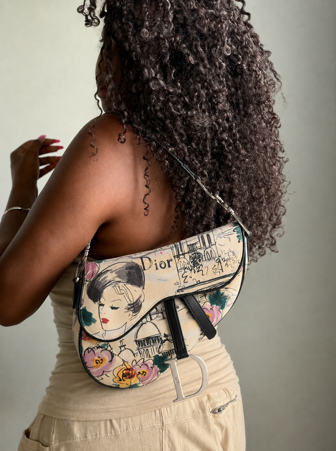 A woman shown from the back wearing a Dior saddle shoulder bag. The bag features a vintage-style cream-colored background with a floral illustration of a woman's face, black script text, and a large silver 'Dior' 'CD' logo charm.